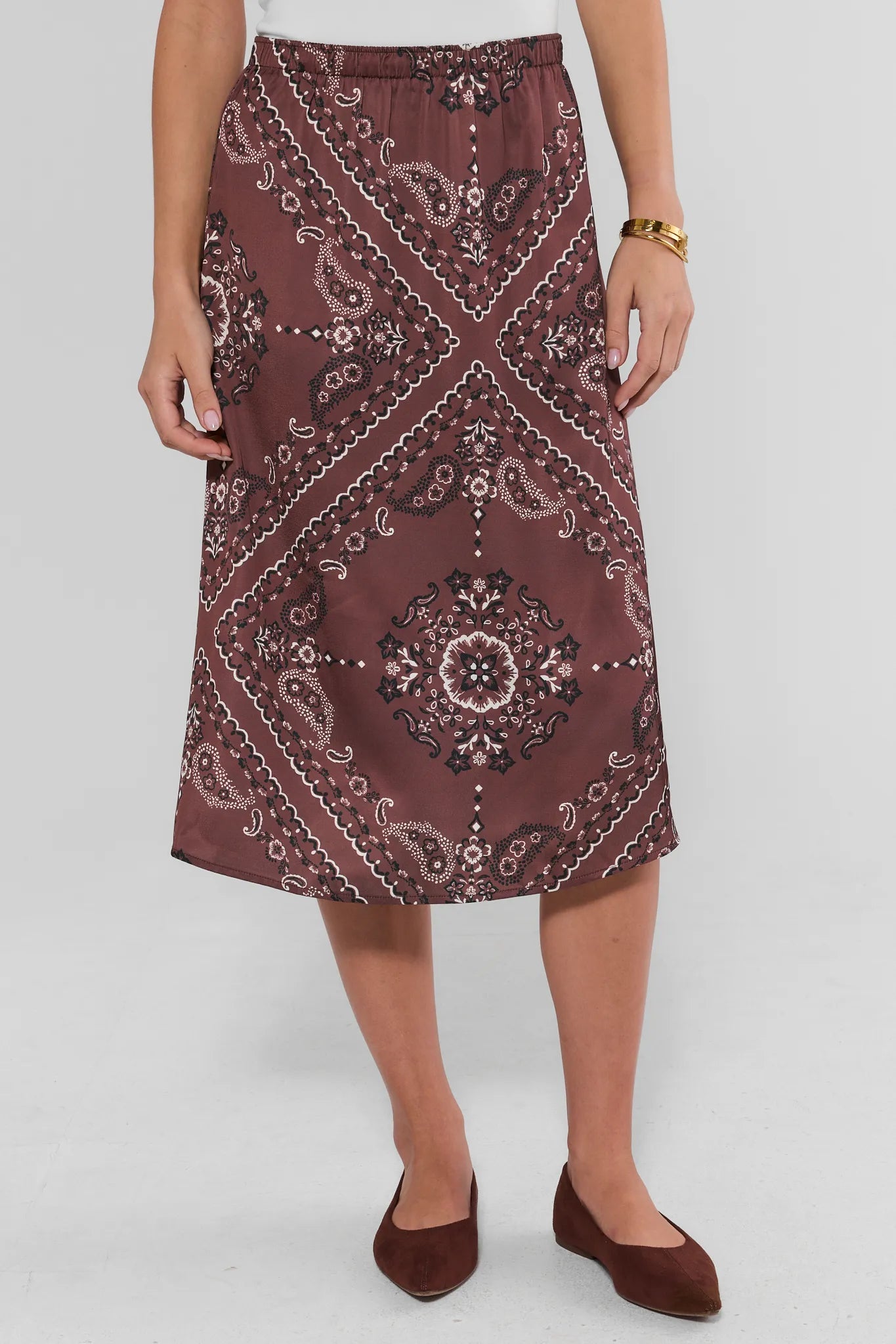 A woman wearing a knee-length Brown Bandana Print Midi Skirt with a white paisley and medallion bandana print, paired with brown flats against a light gray studio background.