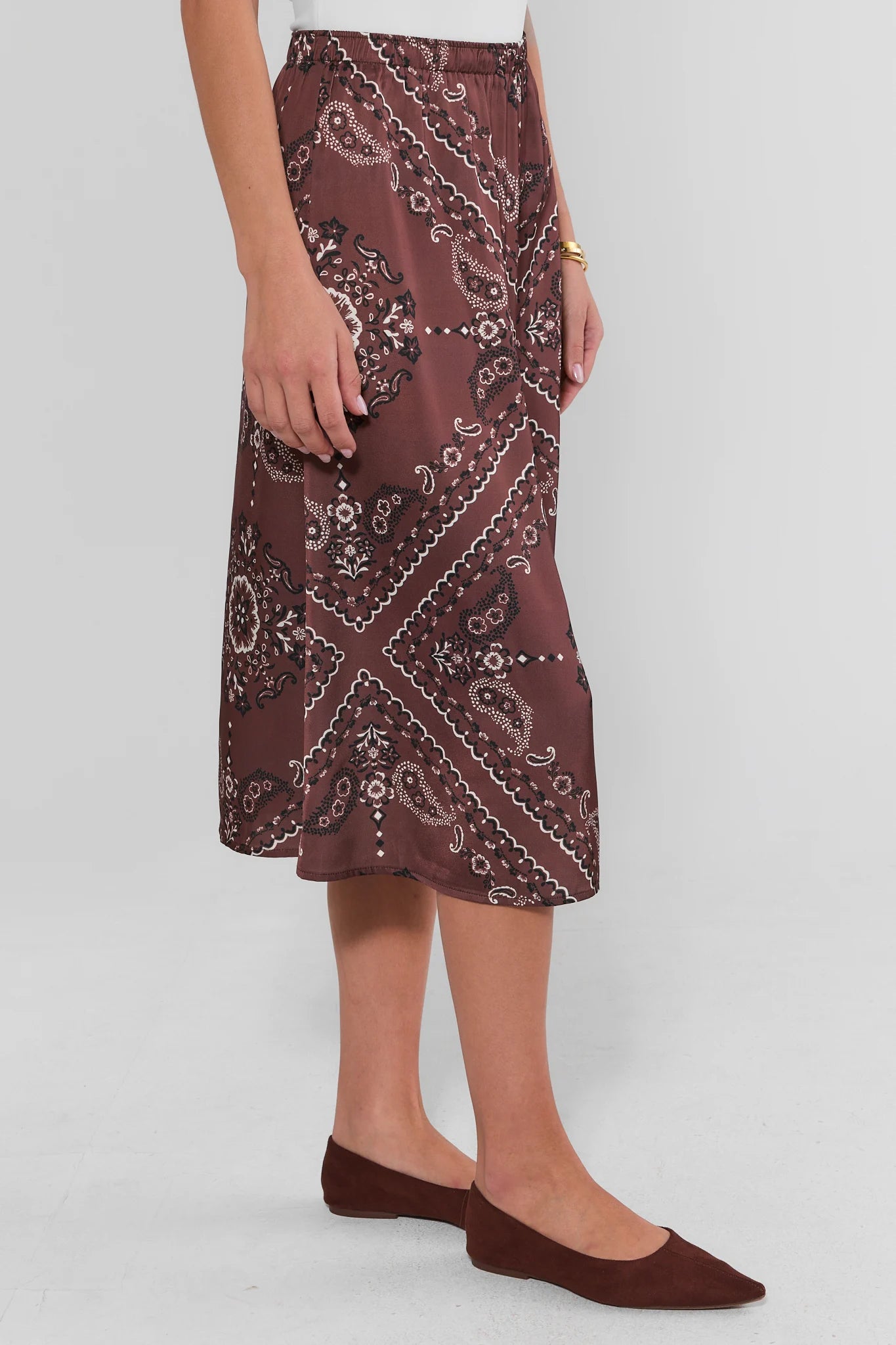 A model wearing the Brown Bandana Print Midi Skirt with a white paisley and bandana print, paired with matching brown flats, shown from the waist down against a light gray background.