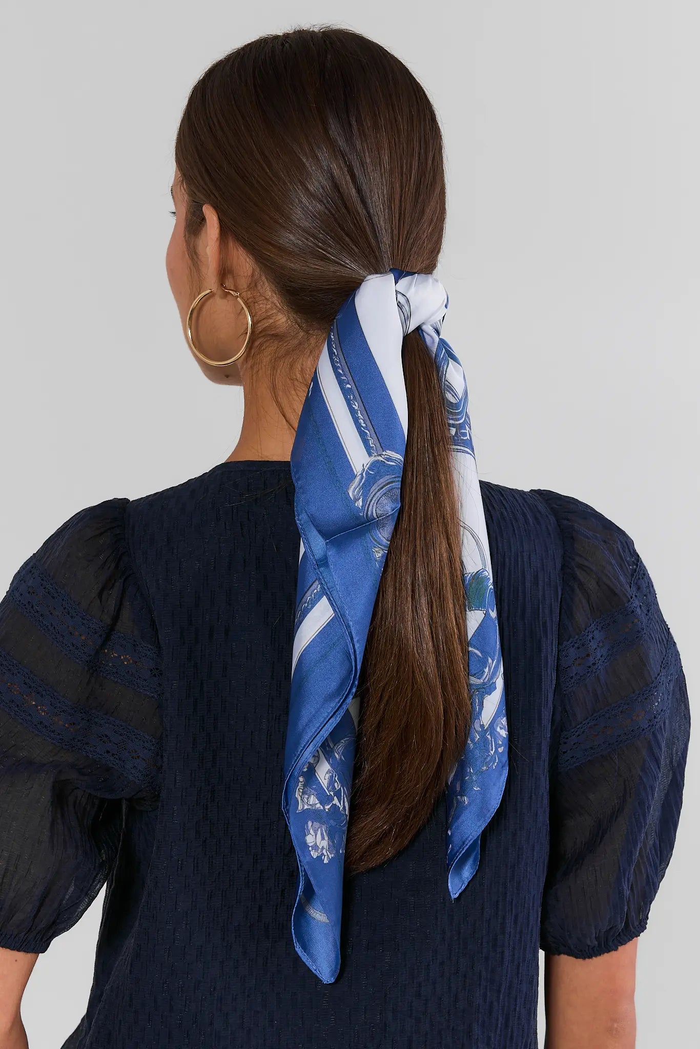 A woman shown from the back wearing a navy blue textured blouse and gold hoop earrings, with her brown hair tied back in a ponytail using the blue chain link print scarf