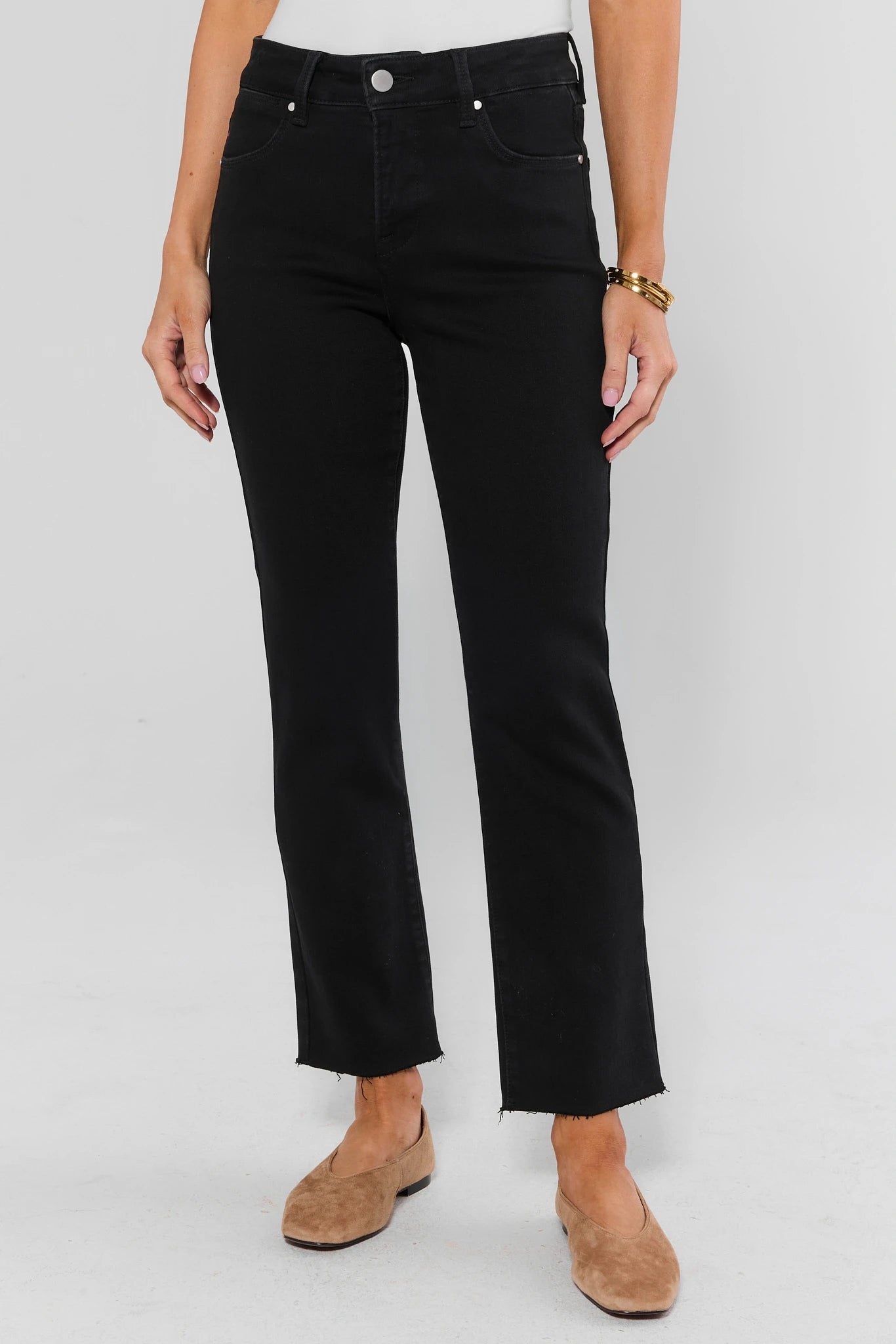 A person wearing black cropped straight-leg jeans with a silver button closure, paired with tan suede flat shoes, against a light gray studio background.