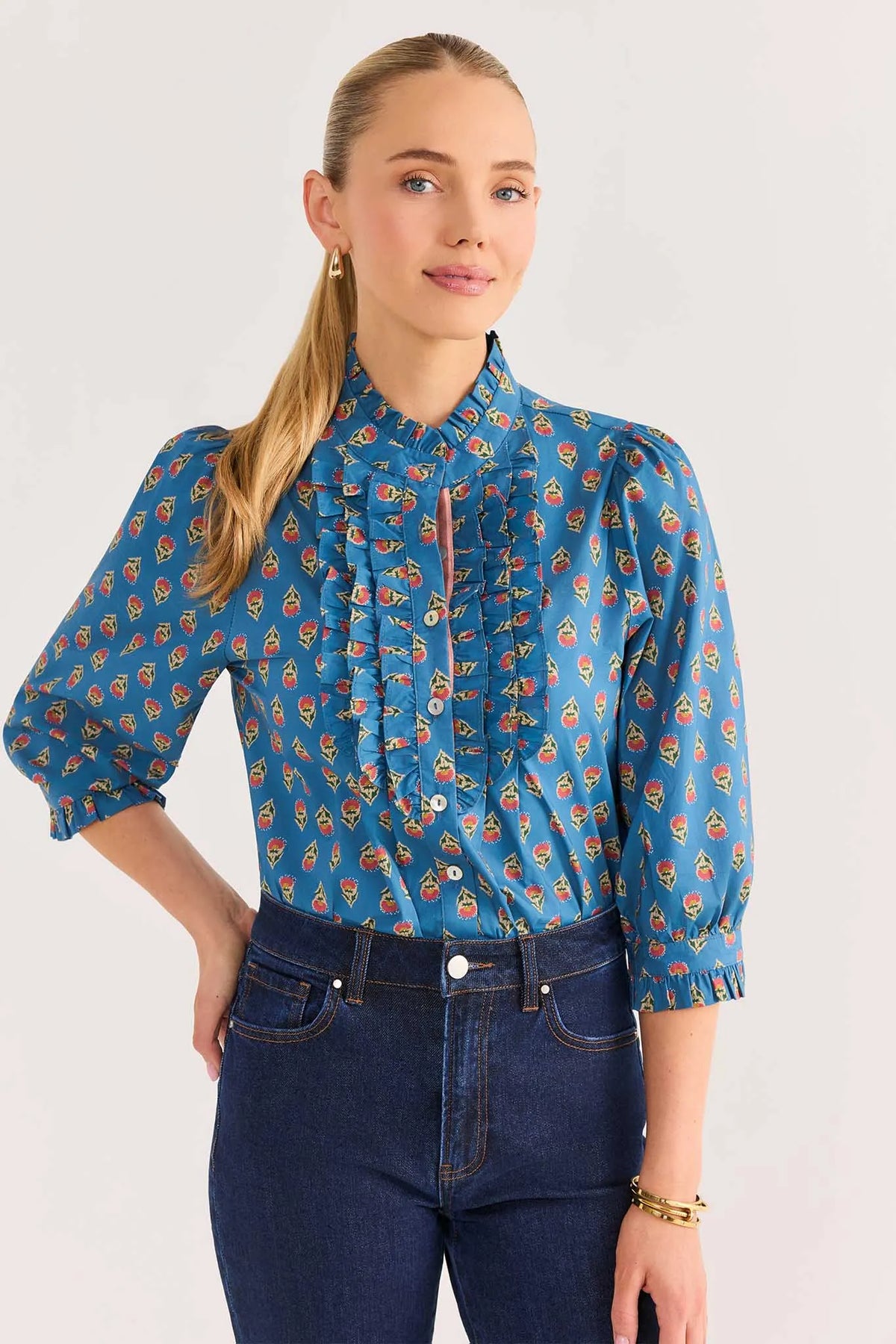 A woman modeling a blue long-sleeve blouse with an allover paisley pattern in orange, yellow, and white, paired with dark blue jeans against a plain light gray background.