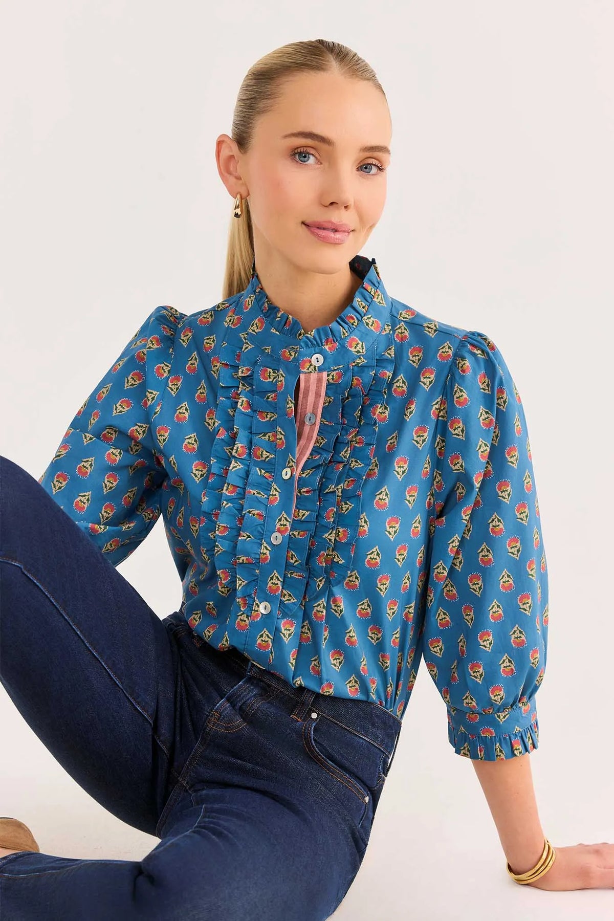 A woman wearing a blue long-sleeve blouse with a colorful cartoon pattern and dark blue jeans, sitting against a plain white background.