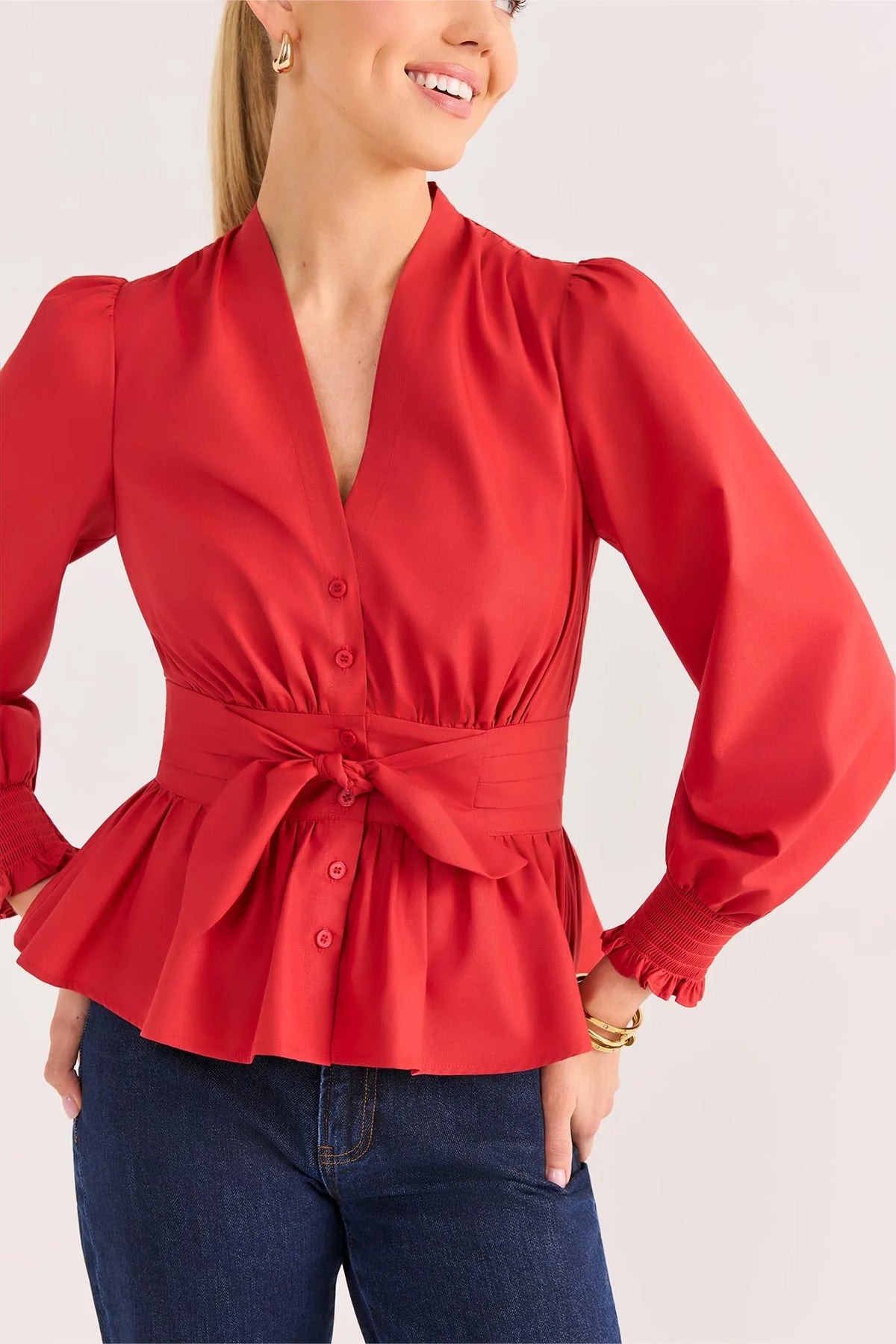 A woman wearing a bright red long-sleeve blouse with a V-neck, button front, and tied waist sash, paired with dark blue jeans against a light neutral background.