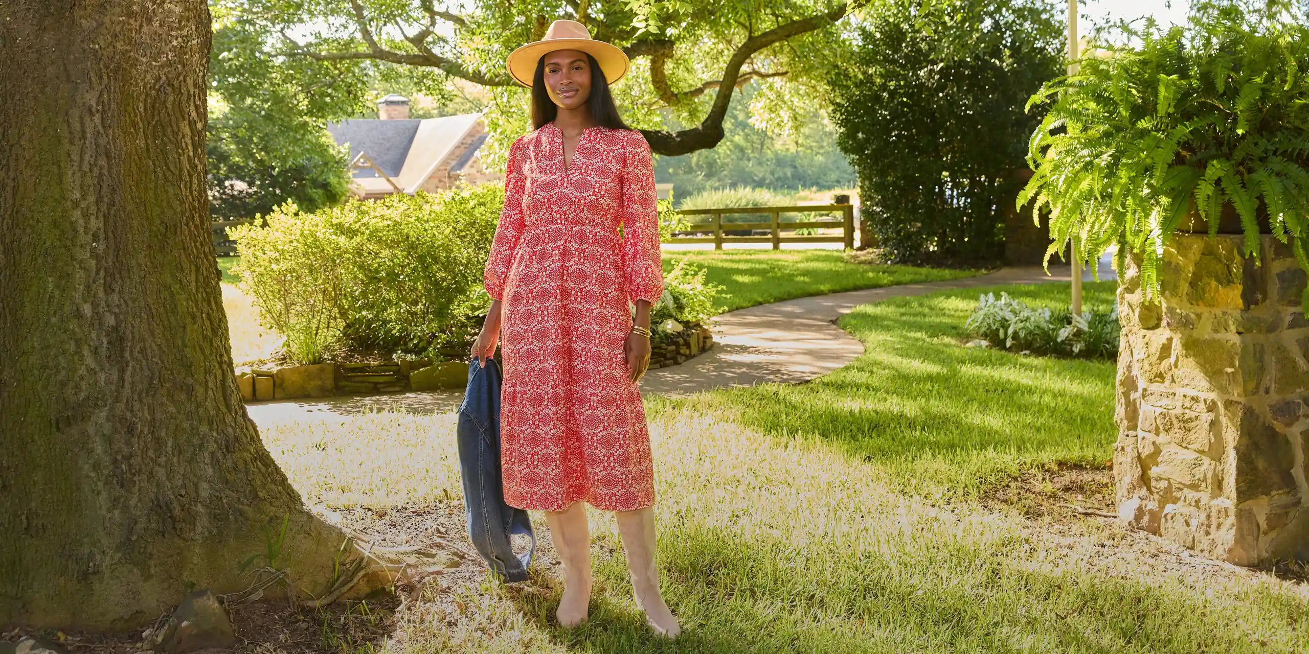 A woman stands outdoors in a garden wearing a pink patterned midi dress, a wide-brim straw hat, and holding a blue garment. The background features a stone wall, lush greenery, and a tree trunk on the left.