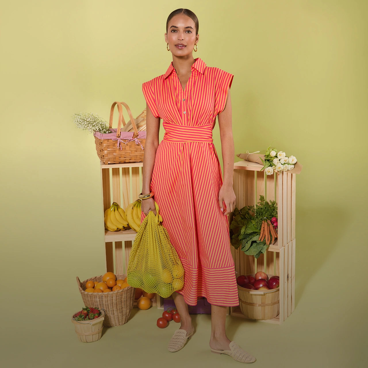 Woman in a red striped dress holding a yellow bag with fruits and vegetables against a green background