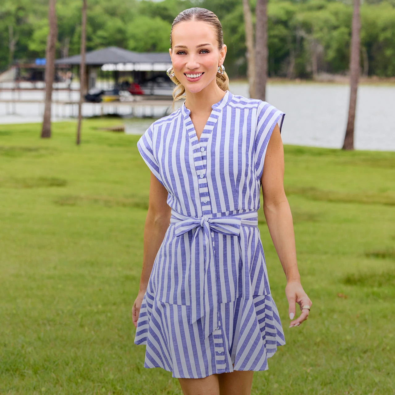 Woman wearing a blue and white striped dress standing in a grassy area with trees and a lake in the background. desktop