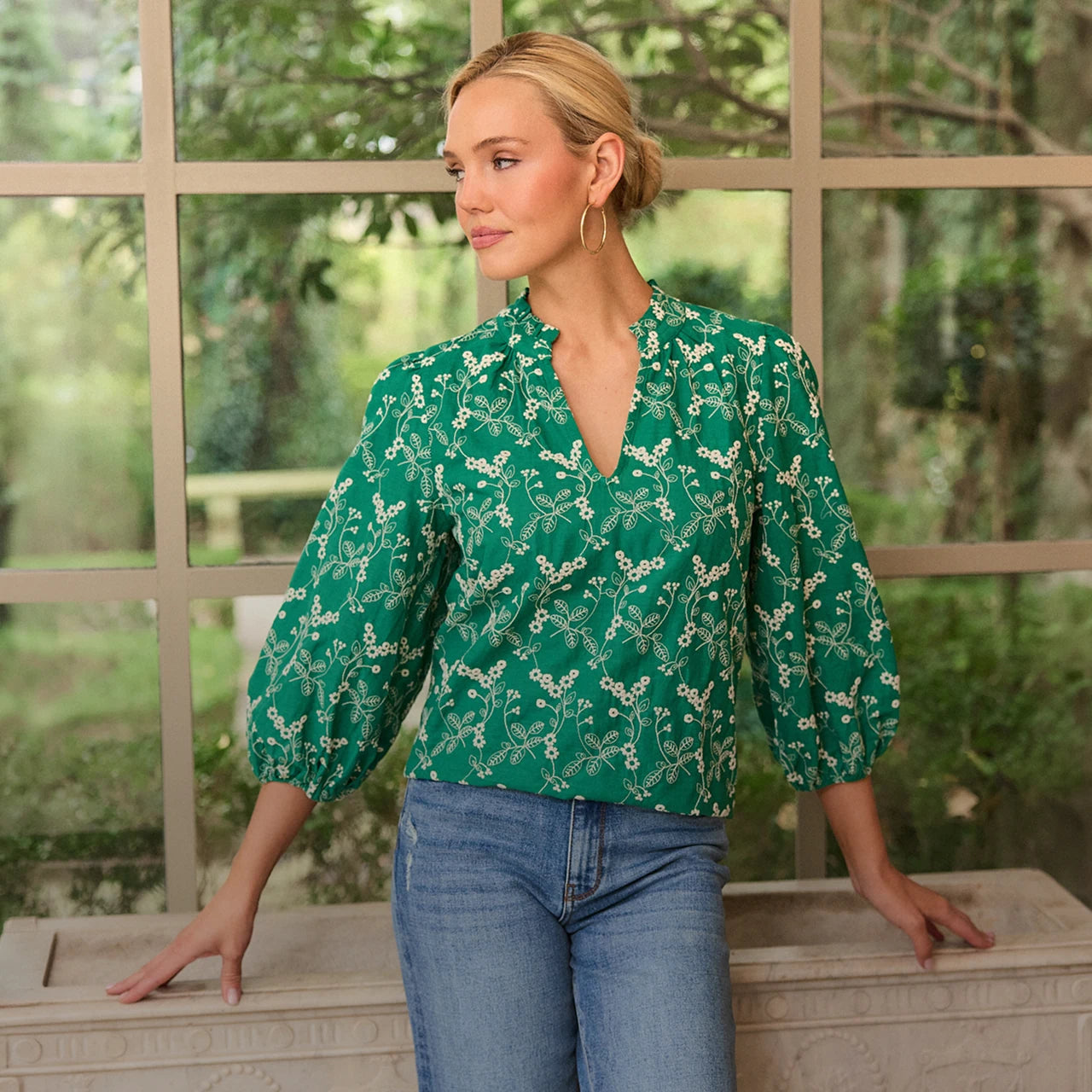 Woman wearing a green floral blouse and blue jeans standing in front of a window with a garden view.