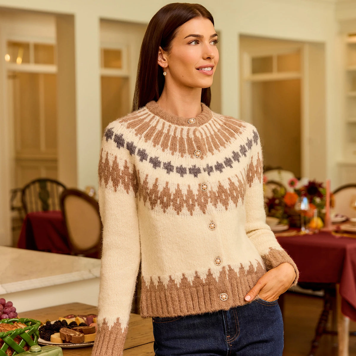 A woman wearing a cream and brown Fair Isle knit cardigan sweater with blue jeans, standing in a warmly lit dining room setting.