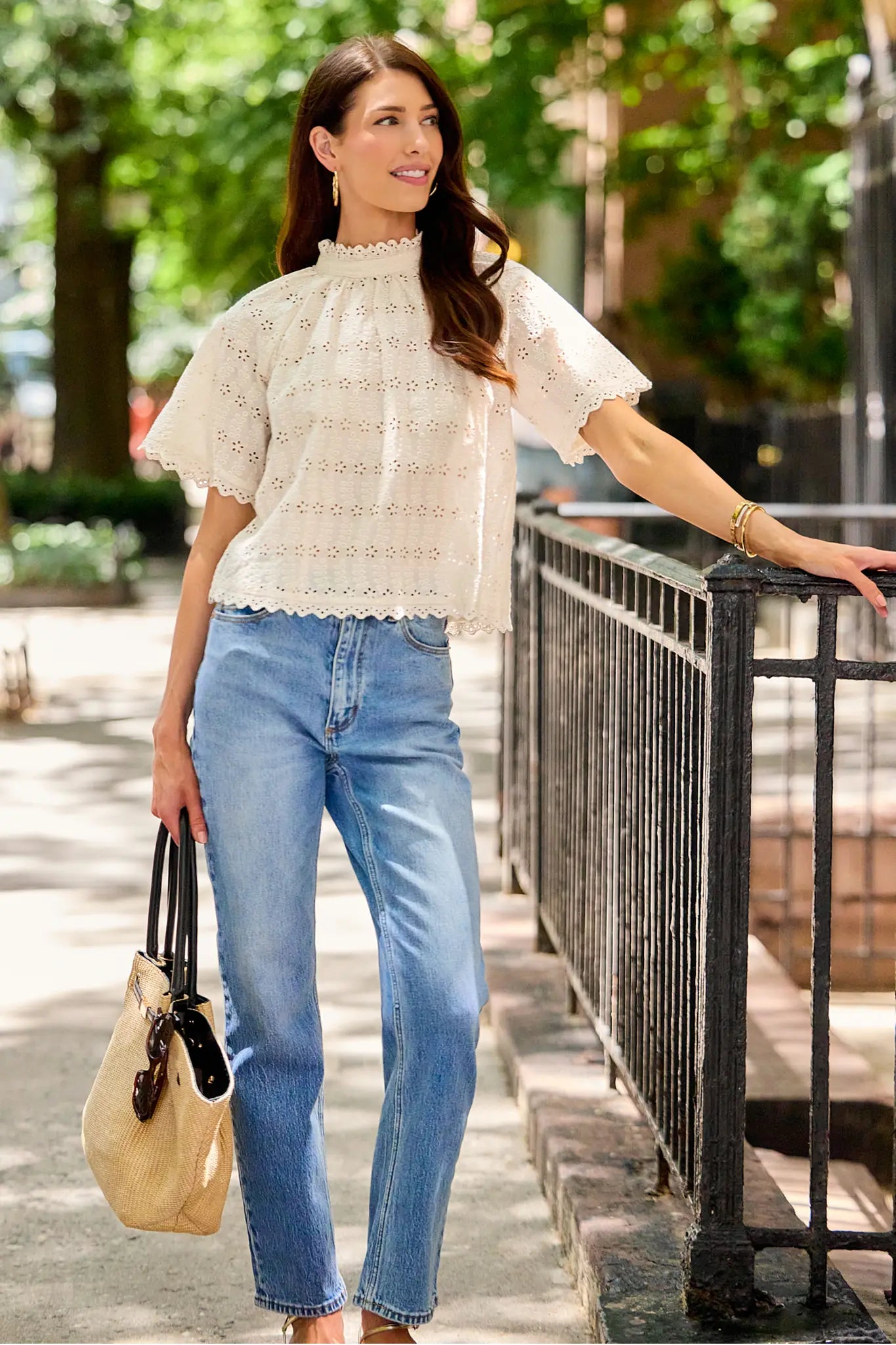 A woman stands outdoors on a sidewalk next to a black metal railing, wearing a cream eyelet short-sleeve blouse and light blue jeans, holding a beige handbag.