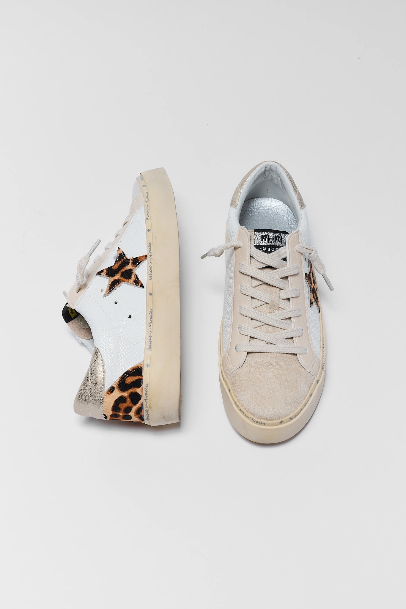 A pair of low-top sneakers shown from the side and front angles on a light gray background. The shoes feature a white leather upper with leopard print overlays on the heel and toe, beige suede-like panels, and beige laces.