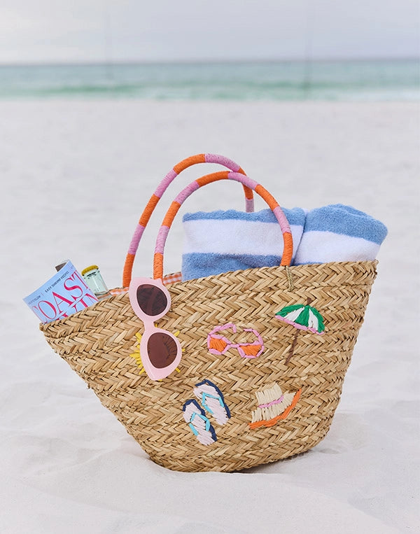 Woven beach bag with sunglasses, towel, and books on a light background. nav