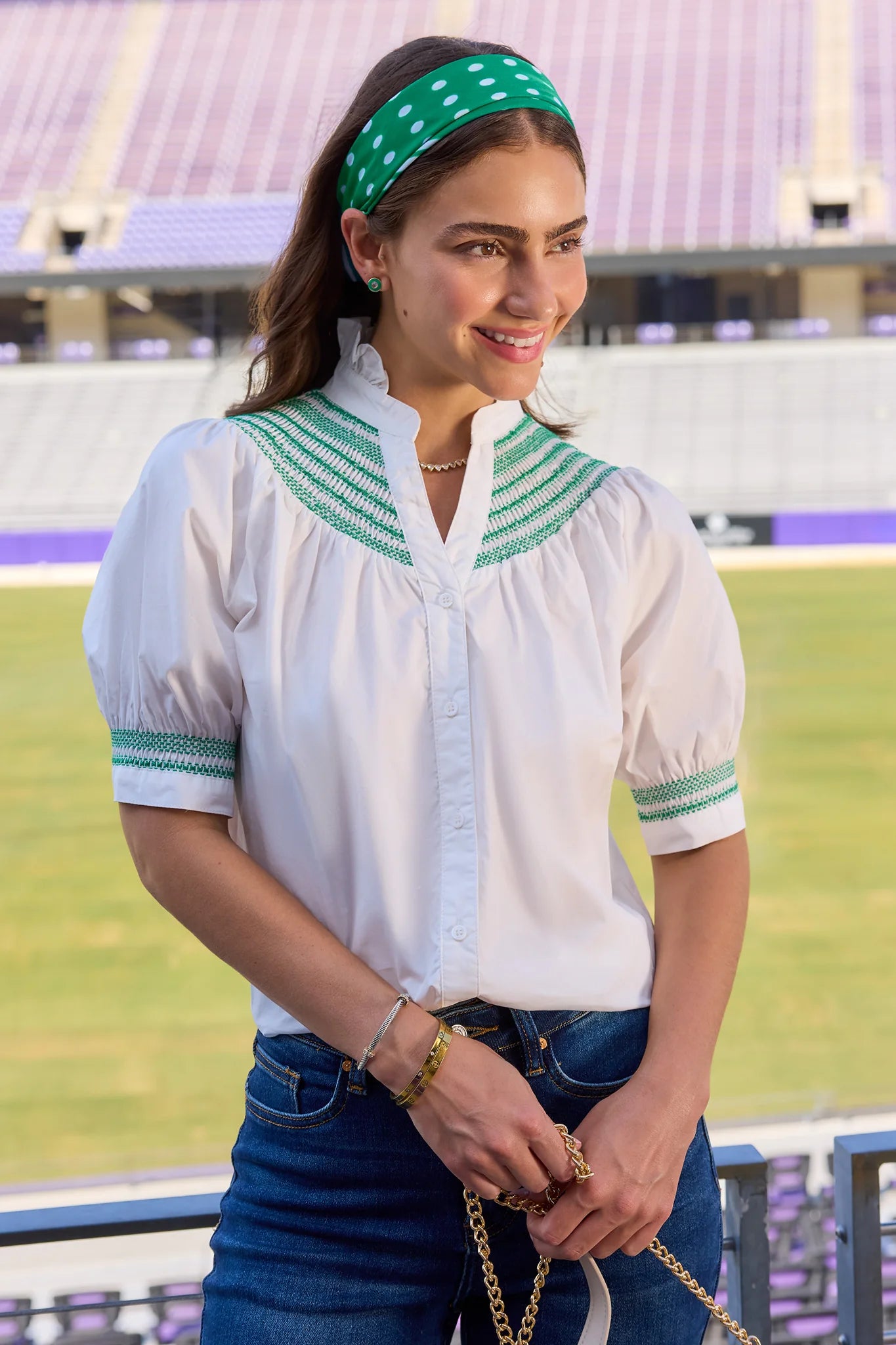 A woman wearing a white short-sleeve blouse with green embroidered trim at the collar and cuffs, paired with a green polka-dot headband and blue jeans, standing in a stadium.