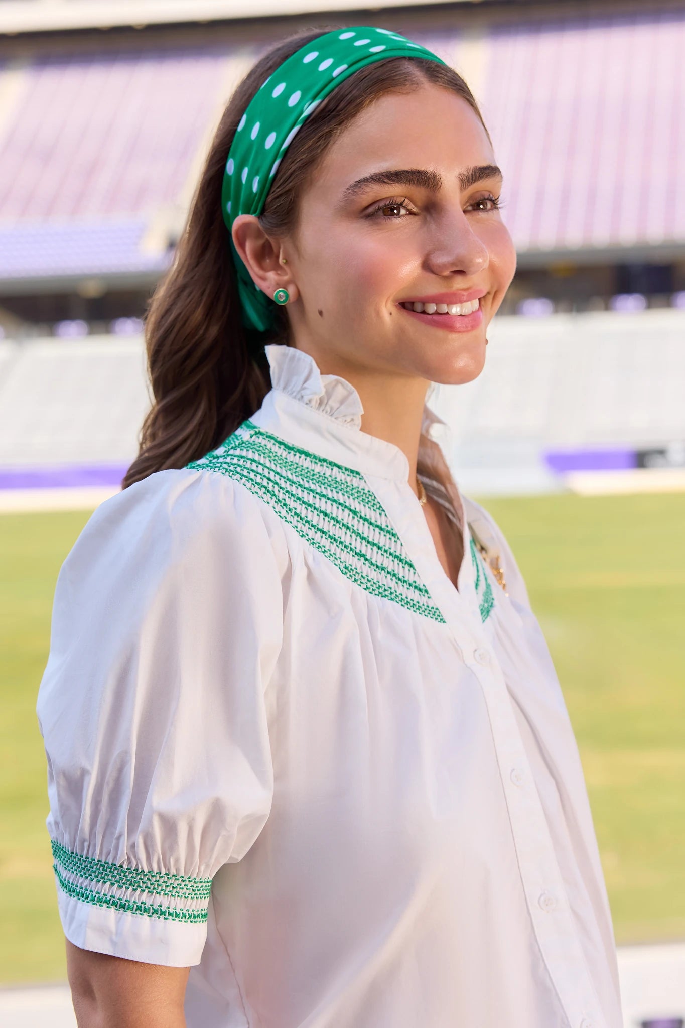 A woman wearing a white blouse with green embroidered trim and a green polka-dot headband, photographed outdoors in front of stadium seating.
