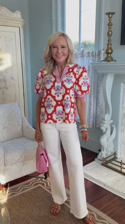 woman wearing mia floral top with white jeans standing inside with blue wall background